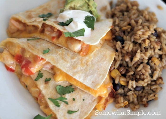 stack of cheesy shrimp quesadillas on a white plate with sour cream and avocado on top and a side of rice