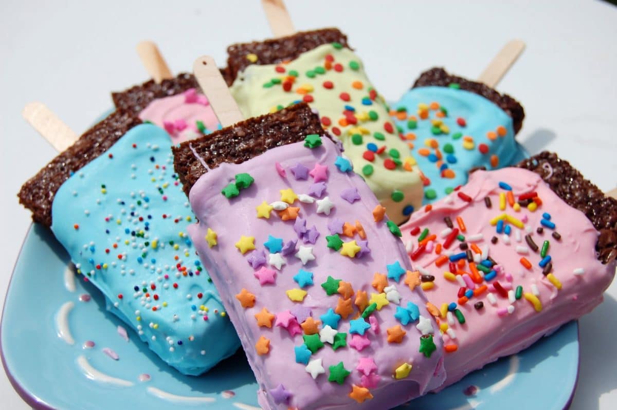 a blue plate filled with several popsicle brownies. Each brownie is covered in purple, pink, blue or green icing and topped with various multi colored sprrinkes.