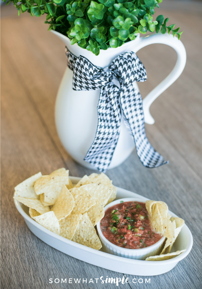 a dish of homemade salsa in a serving bowl filled with tortilla chips sitting on a table next to a potted plant.