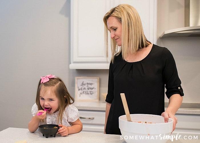 an attractive blonde mom looking at her young daughter eating salsa straight from the bowl with a spoon.