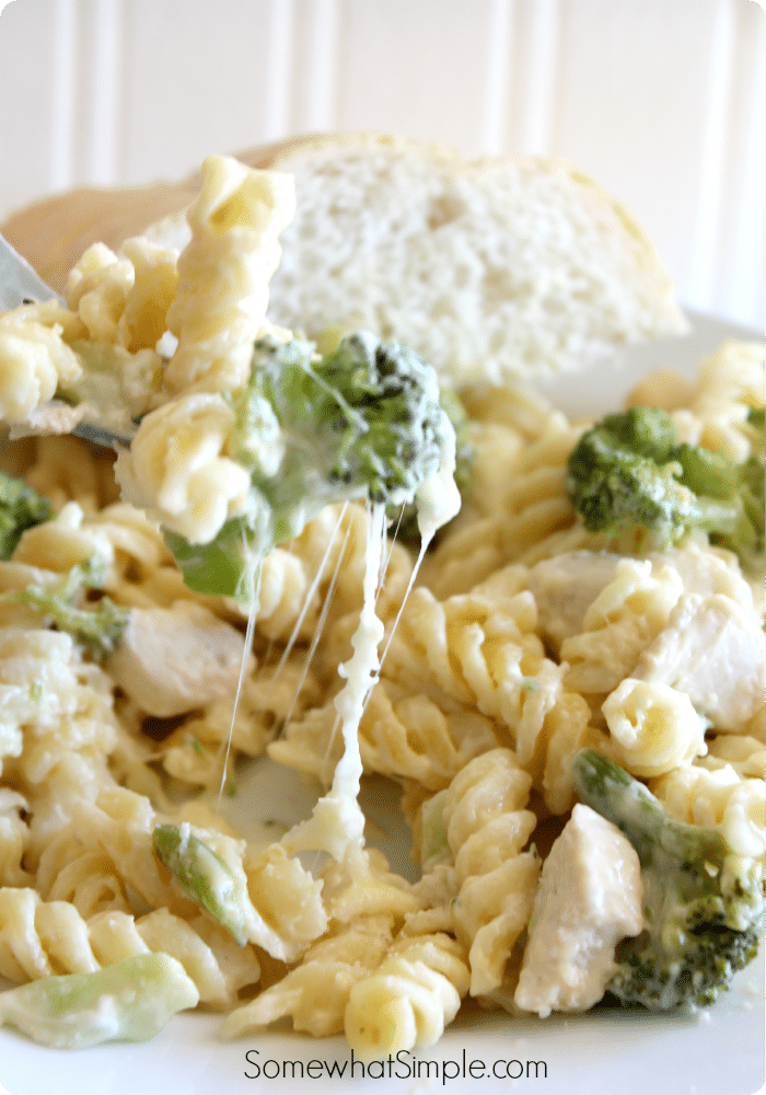 a fork lifting up a few pasta noodles and a piece of broccoli with cheese stretching from the plate of chicken pasta casserole. A slice of bread is in the background.