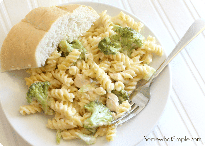 a white plate filled with cheesy chicken broccoli pasta. A slice of bread and a fork are on the plate next to the baked pasta.