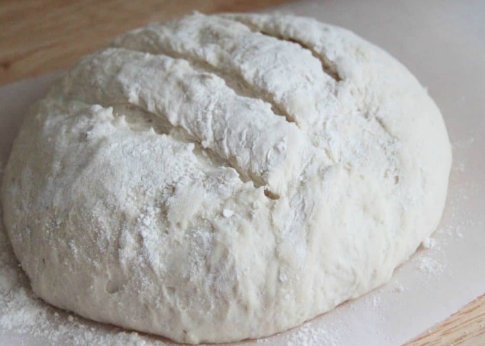 a loaf of uncooked artisan bread on parchment paper with slits cut into the top and flour over the dough