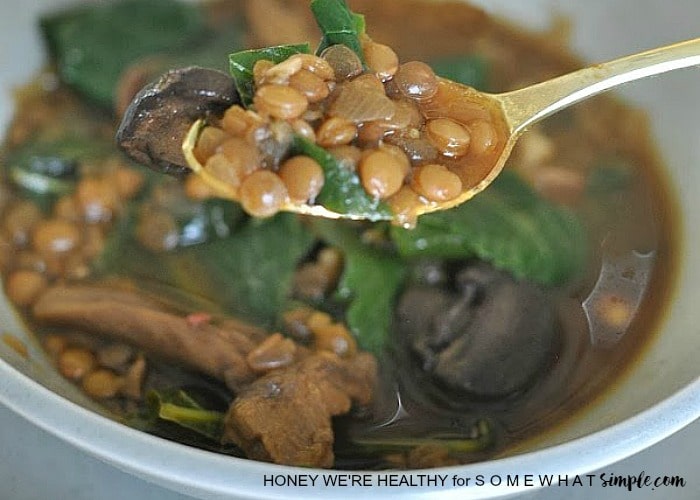 a spoon filled with lentil soup hoovering over a bowl of lentil bean soup with spinach mushrooms and other vegetables