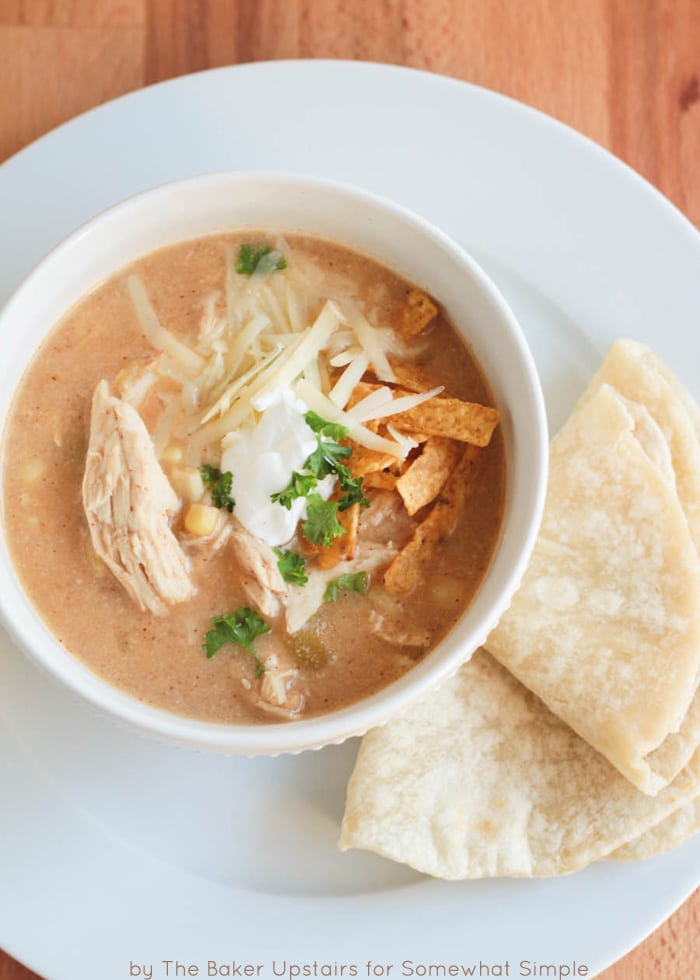 looking down on a white bowl of White bean Chicken Chili that was made in a slow cooker. The chili is topped with shredded cheese, cilantro, sour cream and tortilla strips. The bowl is on a white plate with a tortilla laying on the plate next to the bowl.