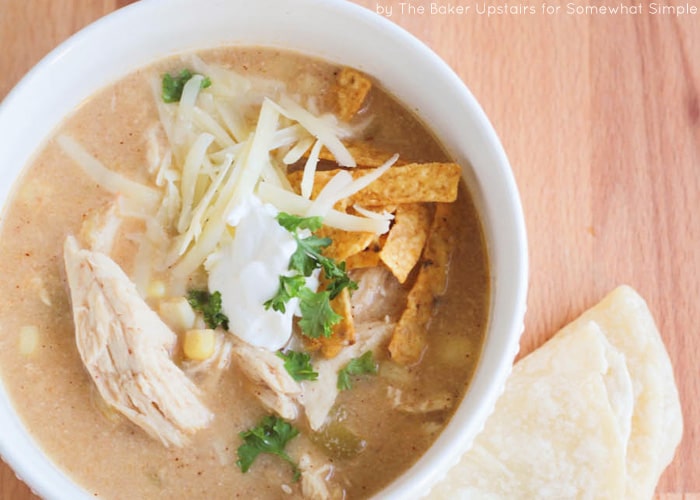 looking down on a white bowl of White Chicken Chili that was made in the crock pot. The chili is topped with shredded cheese, cilantro, sour cream and tortilla strips with a tortilla laying on the table next to the bowl.