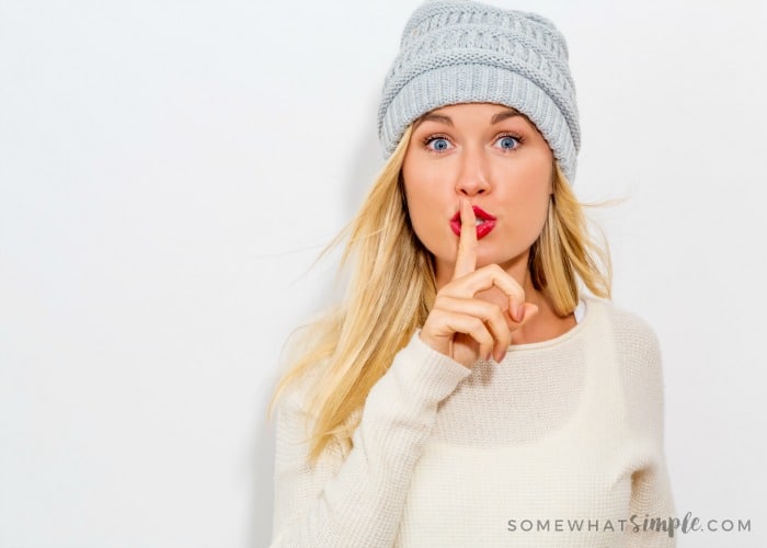 a cute blonde woman with red lipstick wearing a white shirt and grey beanie holding her finger to her lips and mouthing the word shhhh as if she just played an April fools pranks on her husband or boyfriend and is trying to keep it a secret