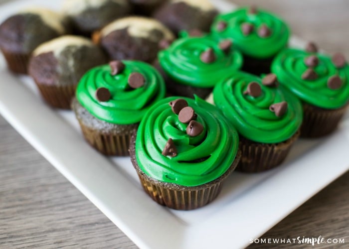 a tray of camo cupcakes on a white tray. half of the army cupcakes are topped with a green frosting and chocolate chips