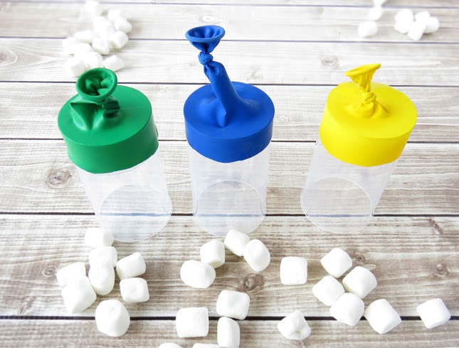 three diy Marshmallow Shooters standing up on a wood table with multiple piles of tine marshmallows around them