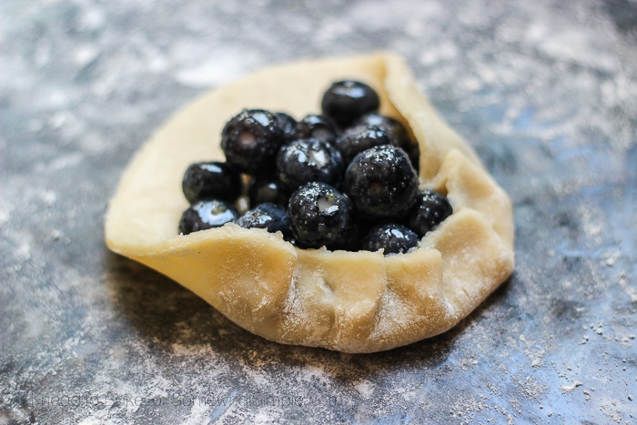 a flat piece of pastry dough being folded up around a pile of blueberries