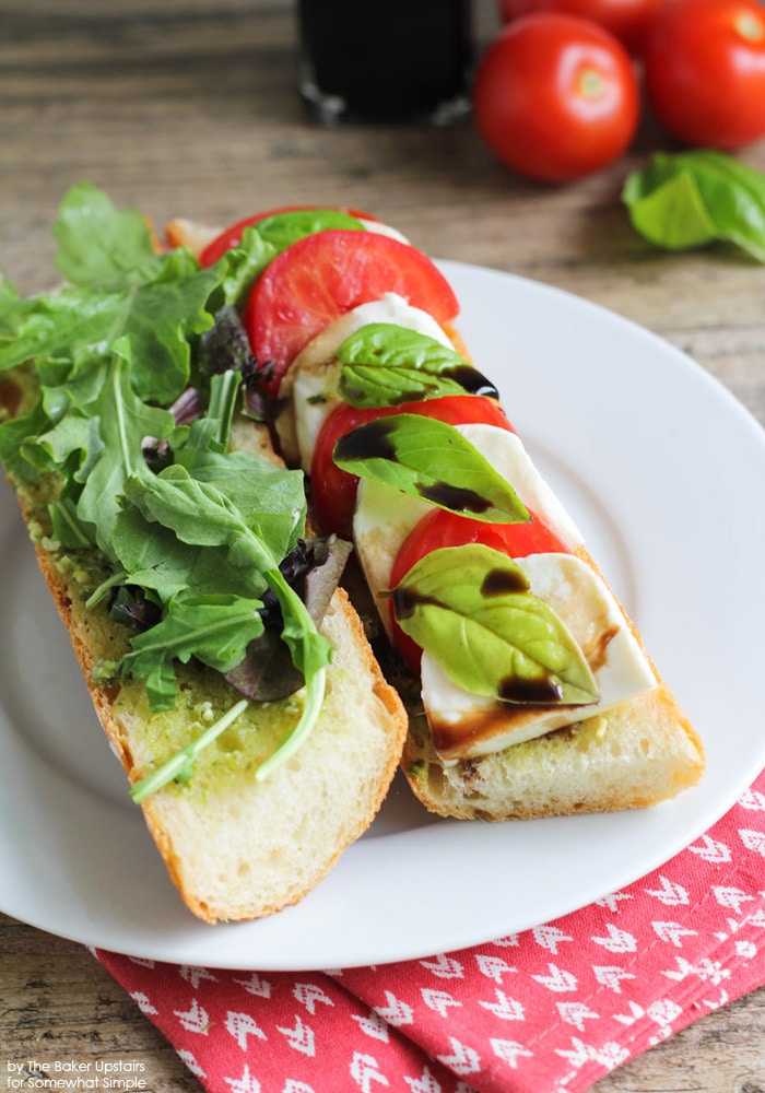 two halves of a Caprese Sandwich on a white plate using this Recipe. One side has lettuce and pesto and the other side has basil, balsamic, tomatoes and mozzarella cheese.