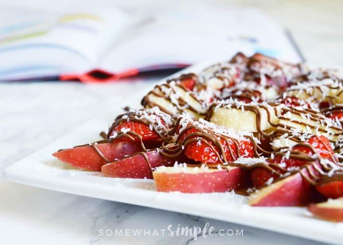 a plate of apple nachos topped with slices of strawberries and bananas, shredded coconut and chocolate drizzled over the top