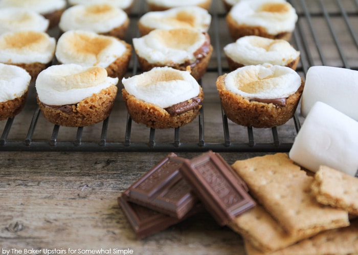 several mini smores bites fresh from the oven sitting on a cooling rack. a few pieces of chocolate, marshmallows and graham crackers are piled in front of the rack.