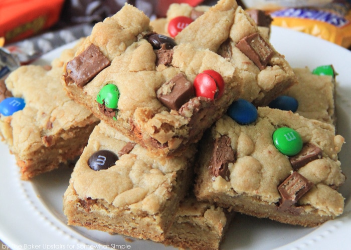 a close up of a white plate filled with Candy Bar Blondies. Each lightly colored brownie is filled with M&Ms or other candy bar pieces.