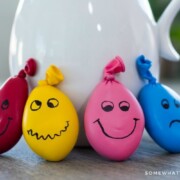 a red, yellow, pink and blue homemade stress ball leaning up against a pot on a table. Each DIY stress ball has a funny face drawn on each one.