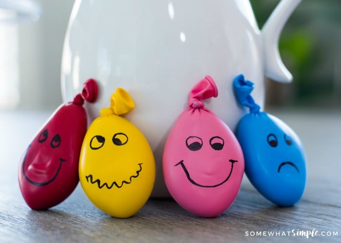 a red, yellow, pink and blue homemade stress ball leaning up against a pot on a table. Each DIY stress ball has a funny face drawn on each one.