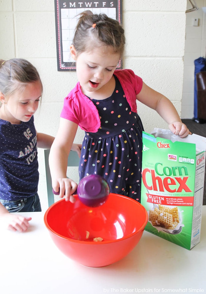 two little girls making Chex Muddy Buddies by pouring ingredients into a bowl with a box of Corn Chex next to the bowl on the counter