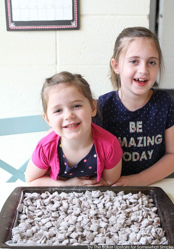 two girls with a baking sheet full of finished Chex mix Muddy Buddies covered in powdered sugar