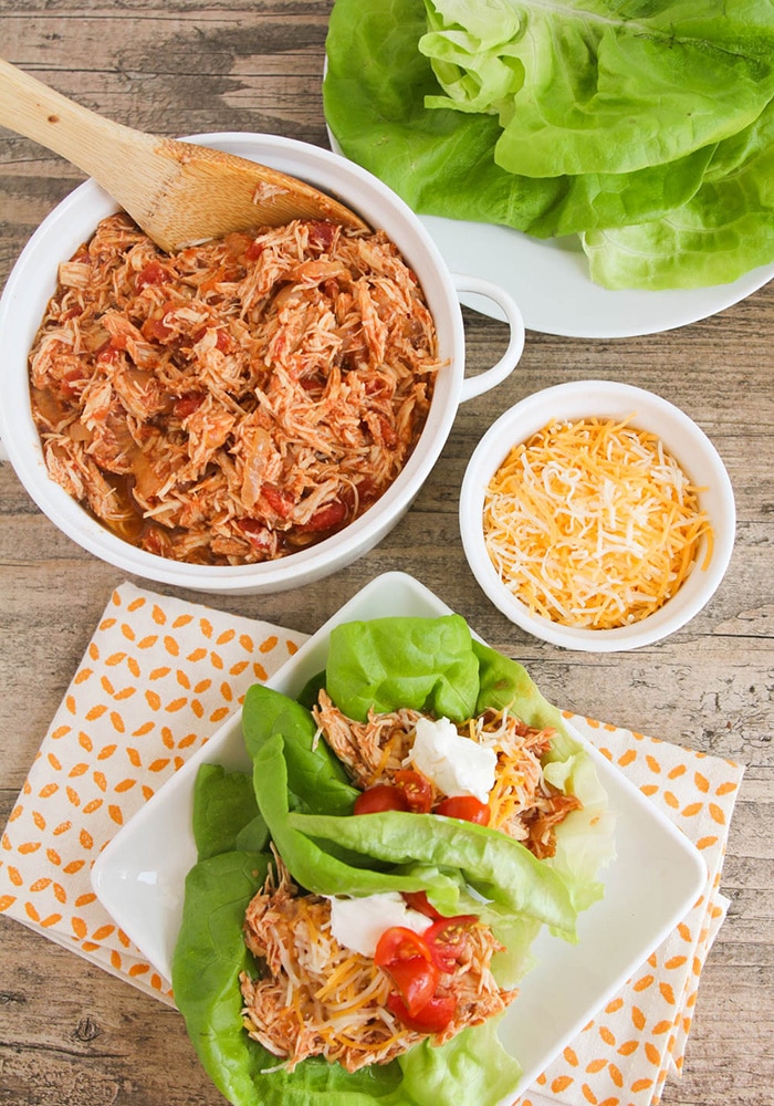 Looking down on a plate with Taco Lettuce Wraps topped with sour cream, tomatoes and cheese. Next to the plate is a bowl of shredded chicken, a small bowl of cheese and a plate of lettuce leaves.