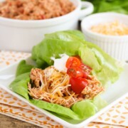 a square white plate with two Chicken Taco Lettuce Wraps on it. Each wrap is topped with shredded cheese cherry tomatoes. Behind the plate is a bowl of shredded chicken and a smaller bowl of shredded cheese.