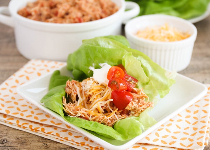 a square white plate with two Chicken Taco Lettuce Wraps on it. Each wrap is topped with shredded cheese cherry tomatoes. Behind the plate is a bowl of shredded chicken and a smaller bowl of shredded cheese.