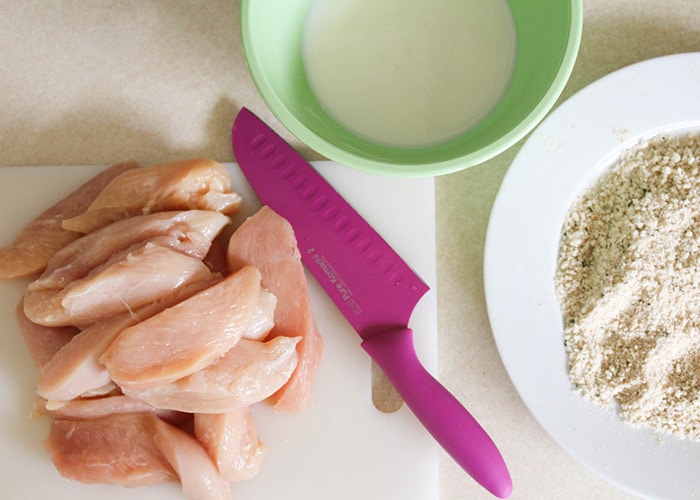 looking down on a cutting board piled high with strips of raw chicken next to a bowl of buttermilk and a plate of bread crumbs