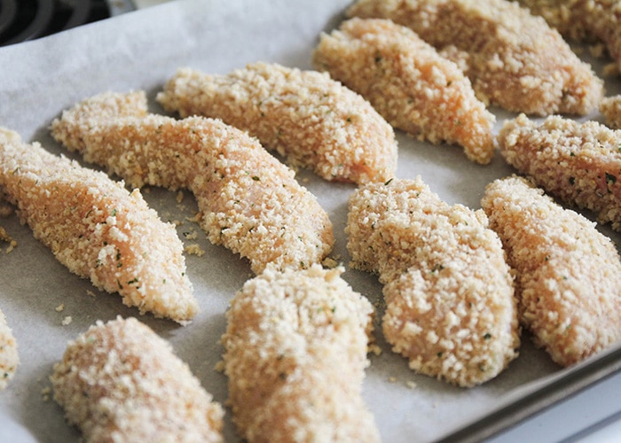 a baking sheet filled with strips of chicken covered in breading ready to go in the oven