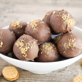 a white bowl filled with nutter butter balls covered in chocolate. Individual nutter butter cookies are scattered on the table around the bowl.
