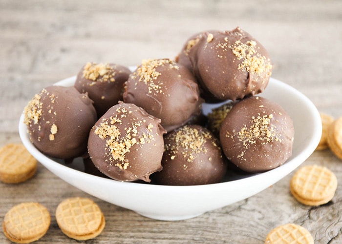 a white bowl filled with nutter butter balls covered in chocolate. Individual nutter butter cookies are scattered on the table around the bowl.