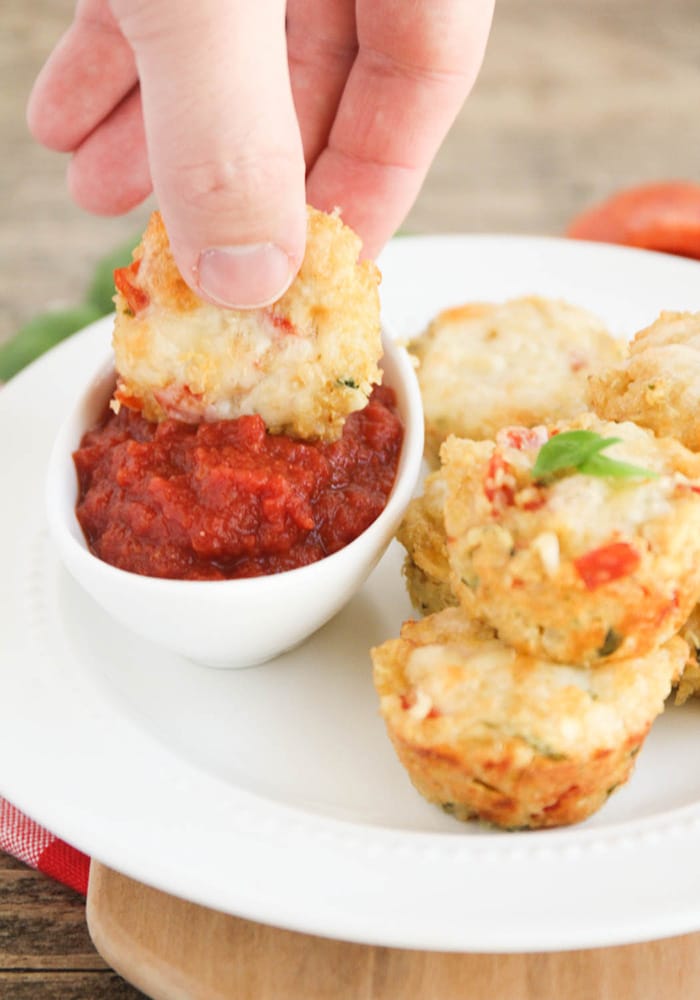 A hand dipping a healthy quinoa pizza bite into a small dish of marinara sauce. Next to the sauce there is a pile of more quinoa bites.