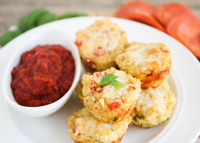 a pile of Quinoa Pizza Bites on a white plate. Next to the pizza bites is a small white sauce dish filled with marinara sauce.