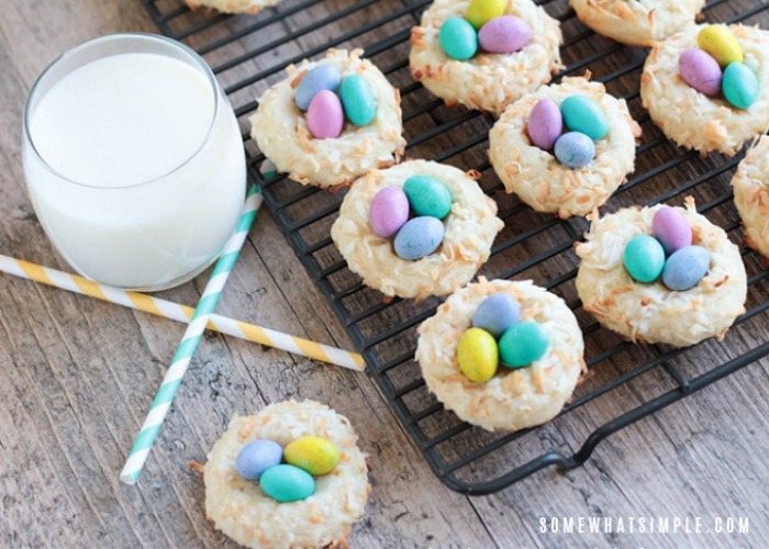 looking down on a black cooling rack filled with Birds Nest Cookies that have three pieces of colored chocolate egg candies in the center of the cookie that look like bird eggs and the cookies are made with coconut so the cookies look like branches are coming out to form a nest. Next to the cooling rack is a glass of milk and below the glass are two pastel colored striped straws that are crossed to look like an X. One bird nest cookie is sitting on the counter below the straws.