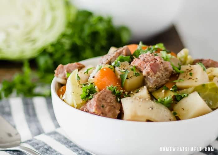 a white bowl of Corned Beef and Cabbage that was made in a slow cooker. Also, in the bowl are slices of onions, potatoes and carrots with chopped parsley on top. A half a head of cabbage is in the background.