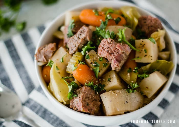 looking down on a white bowl of crock pot Corned Beef and Cabbage that is mixed with carrots celery and potatoes. The bowl is sitting on top of a black and white striped napkin.