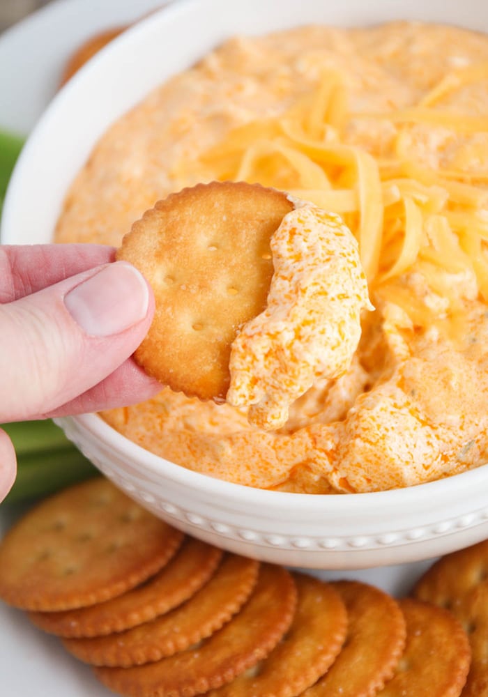 a hand dipping a Ritz cracker into a white bowl filled with this slow cooker buffalo chicken dip.