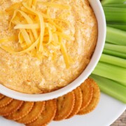 looking down on a white bowl full of this easy buffalo chicken dip recipe that was made in a crock pot and topped with shredded cheese. On the plate next to the bowl are several Ritz crackers and celery sticks