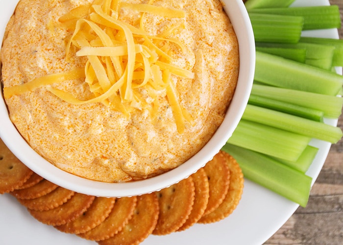 looking down on a white bowl full of this easy buffalo chicken dip recipe that was made in a crock pot and topped with shredded cheese. On the plate next to the bowl are several Ritz crackers and celery sticks