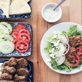 a plate filled with this easy Gyro Salad Recipe next to serving trays filled with the ingredients to make this greek salad as well as a small dish filled with tzatziki dressing