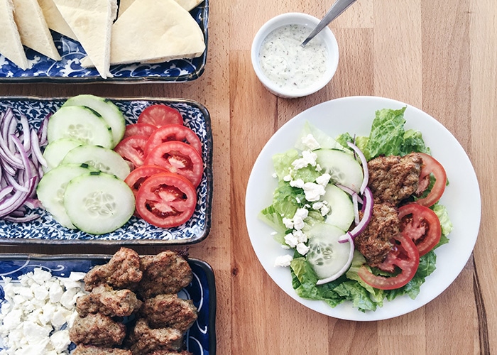 a plate filled with this easy Gyro Salad Recipe next to serving trays filled with the ingredients to make this greek salad as well as a small dish filled with tzatziki dressing
