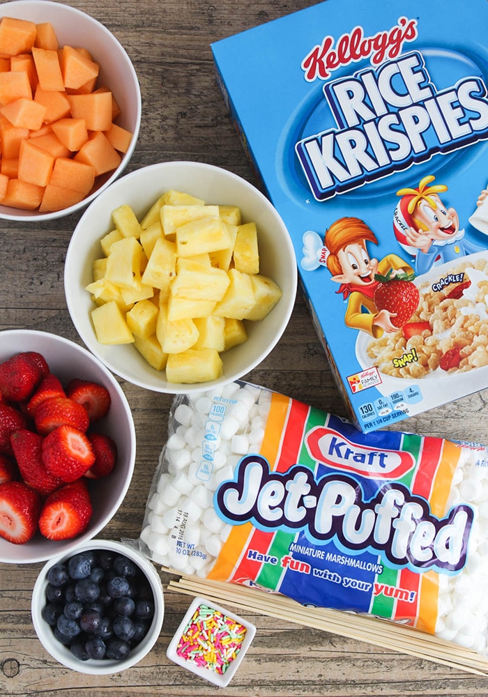 looking down on the ingredients to make these Easter Kebabs. There's a box of rice krispies, a bag of marshmallows, and bowls of cantaloupe, pineapple, strawberries and blueberries.