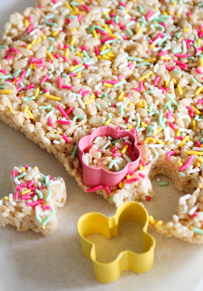 a tray of rice krispie treats covered in pastel colored sprinkles. A pink cookie cutter has be pressed down into the pan to cut out a piece in the shape of a flower.