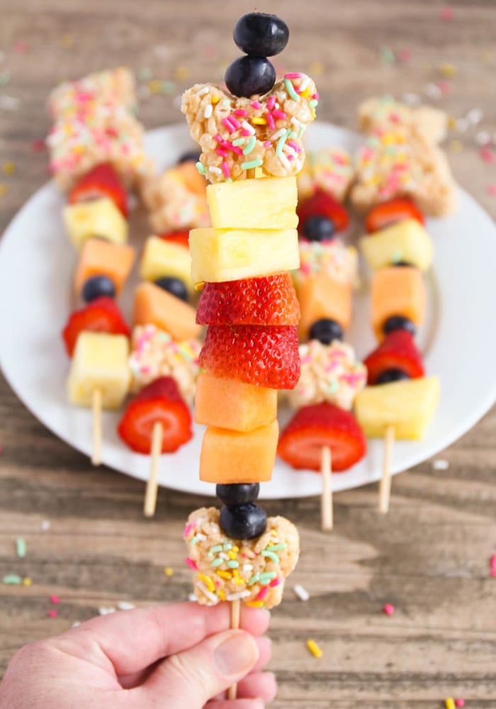 a close up of a Rice Krispies Easter Kebabs being held up with more easter kabobs laying on a white plate in the background. The kebabs are made with sliced pineapple, cantaloupe, strawberries, a few blueberries and pieces of rice krispie treats.