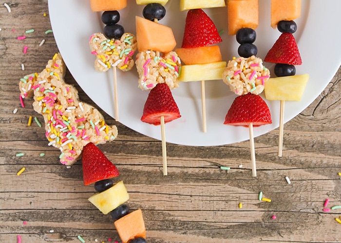 several Easter Kebabs on a white plate with one on the table next to the plate. These Easter snacks are made with rice krispies, pastel colored sprinkles, strawberries, blueberries, cantaloupe and pineapple.