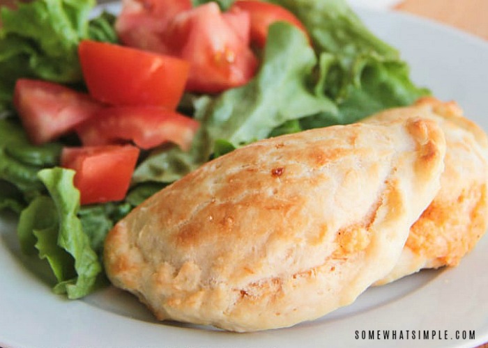 a baked chicken empanada that is golden brown on a white plate next to a green salad topped with tomatoes.