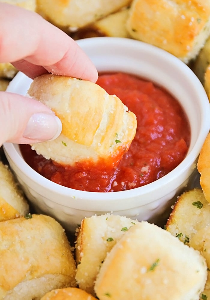 a hand dipping a homemade soft Pretzel Bite into a small dish of marinara sauce. More garlic pretzel bites surround the small bowl.