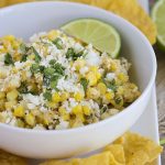 a bowl of Mexican corn salad topped with cilantro and cotija cheese on a serving dish next to tortilla chips