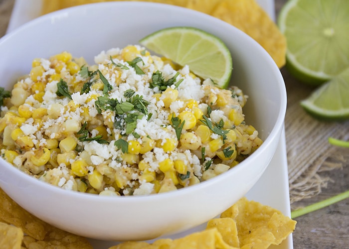 a bowl of Mexican corn salad topped with cilantro and cotija cheese on a serving dish next to tortilla chips