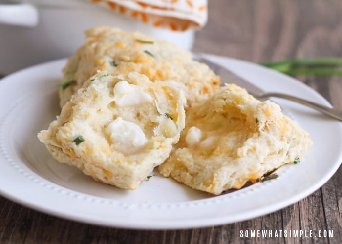 two cheddar biscuits with chives on a plate. One is cut in half with a pad of melted butter on top.