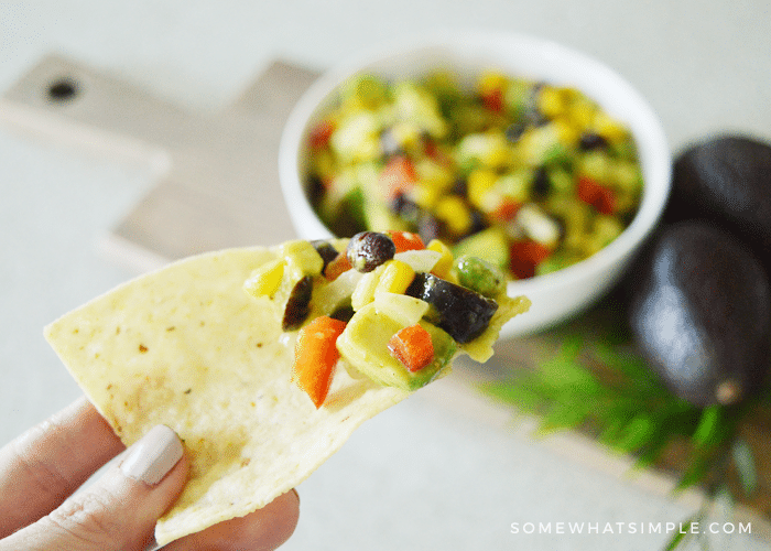 a tortilla chip with a scoop of texas caviar on it. The chip is being held up and in the background below is a bowl filled with texas caviar and two whole avocados.