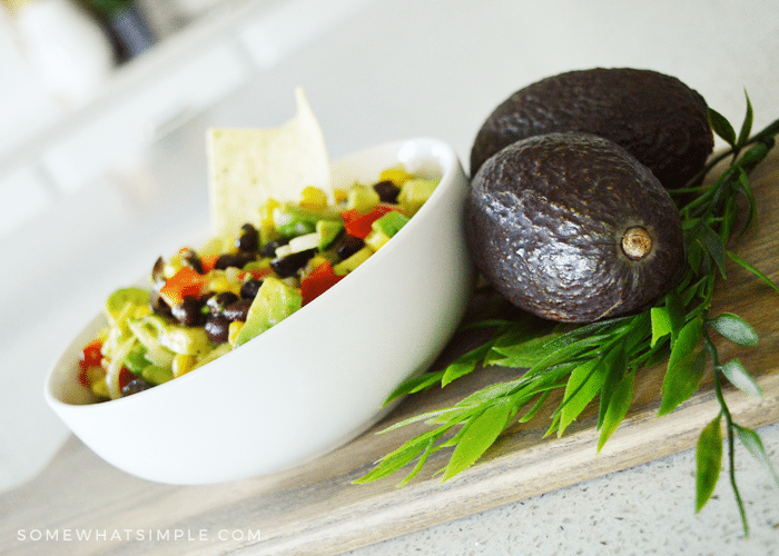 a white bowl filled with texas caviar that's made with black beans, avocado, corn and tomatoes. Next to the bowl are two whole avocados.